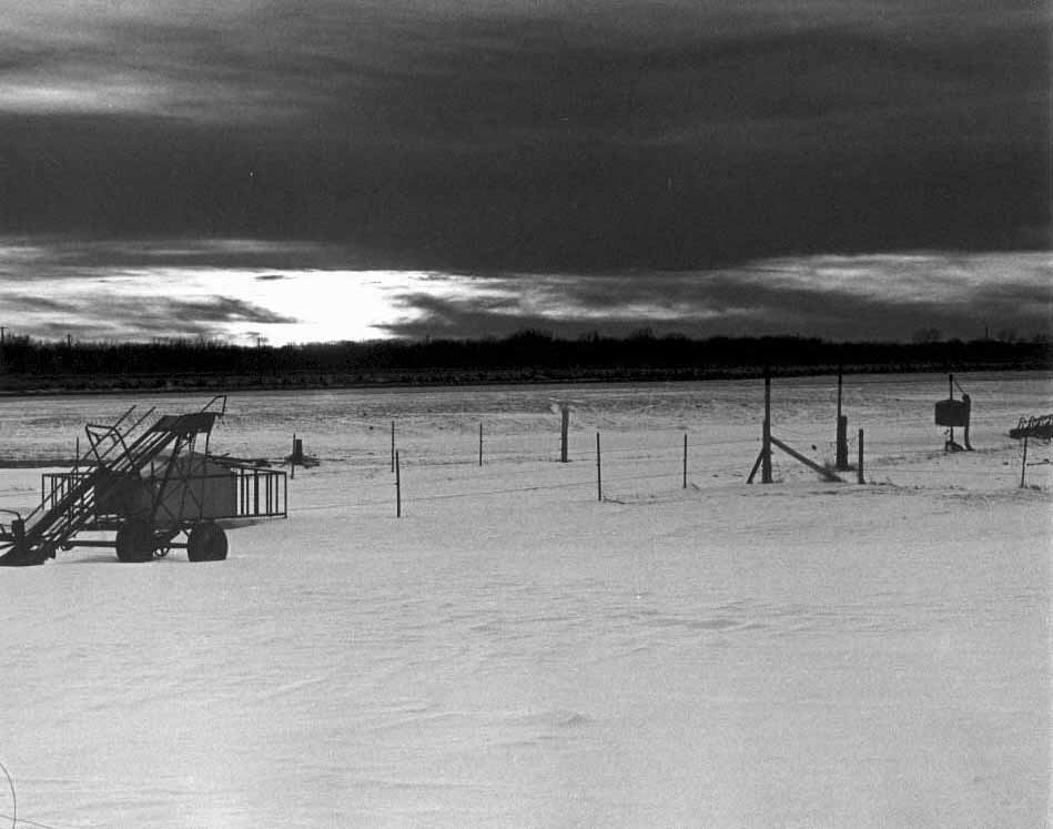 Sun setting on Iowa snow and farmland with deserted equipment in foreground in late February.